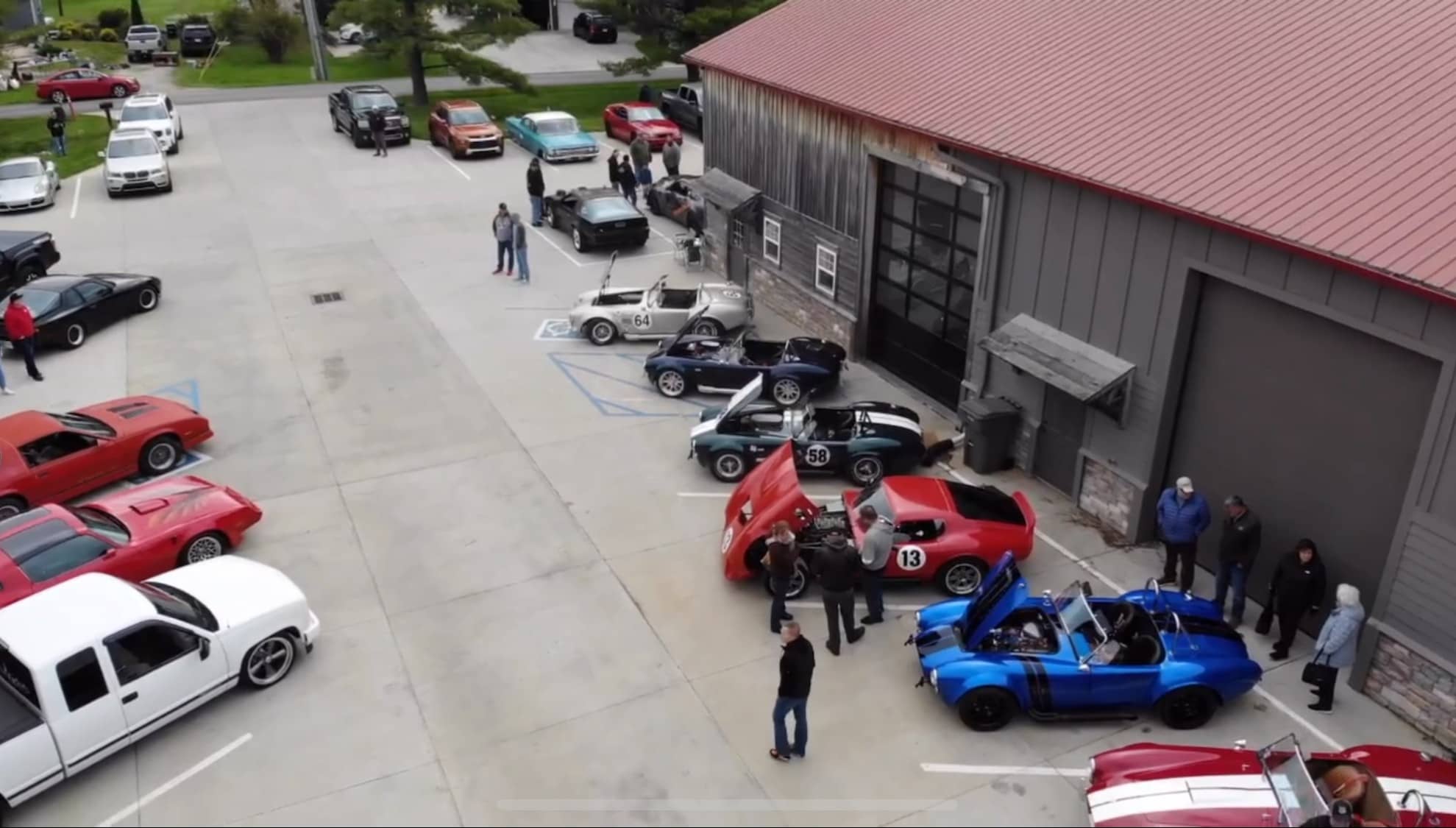 Aerial view of a car show featuring Cobra replicas, a red Daytona Coupe #13, classic muscle cars, and spectators outside a large building with a red roof.