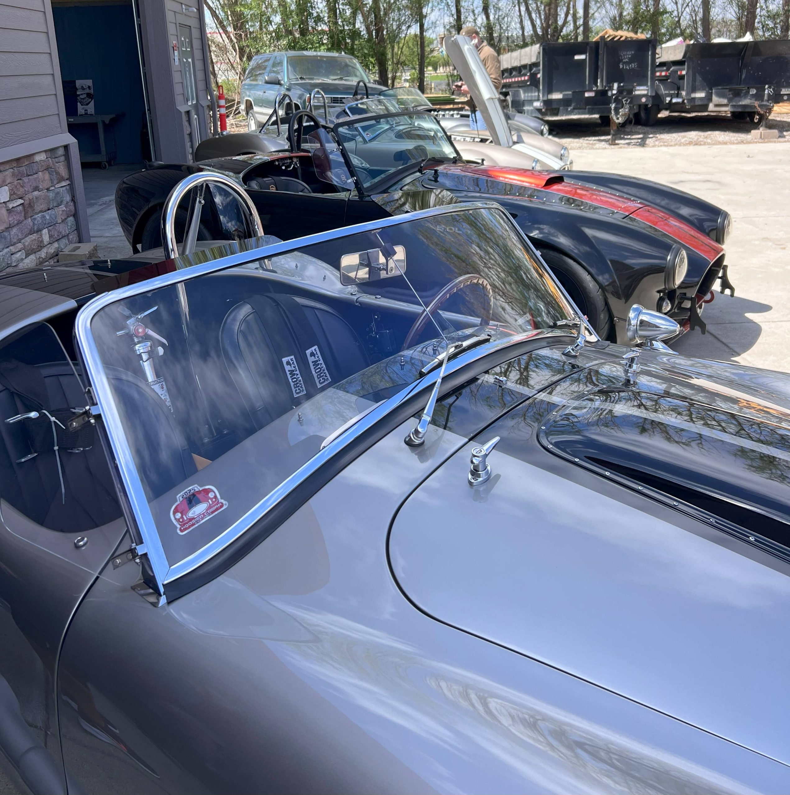 Lineup of Cobra-style roadsters at a car event, including a black model with red racing stripes and a visible roll bar.