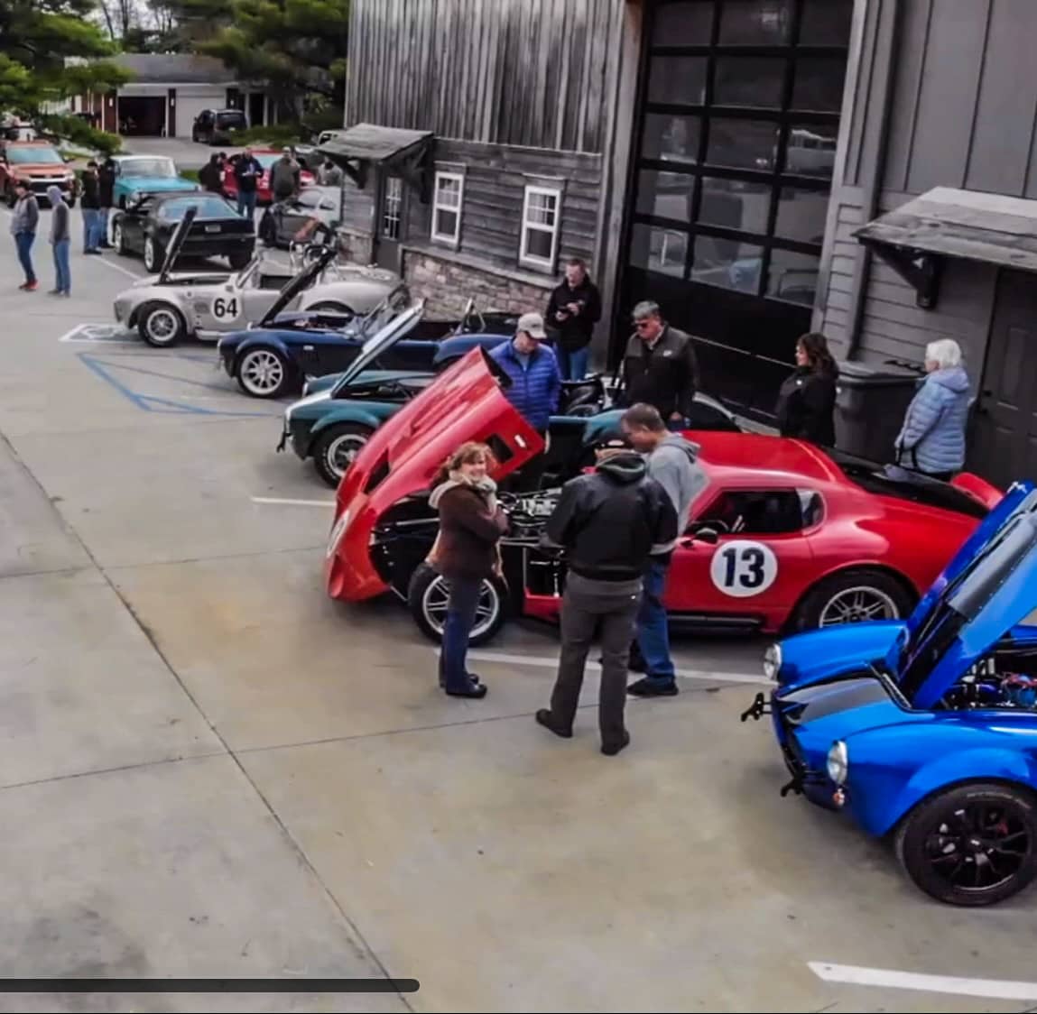 Overhead view of a car show lineup featuring Cobra-style roadsters, a red Daytona Coupe with number 13, and multiple classic and custom cars outside a garage.