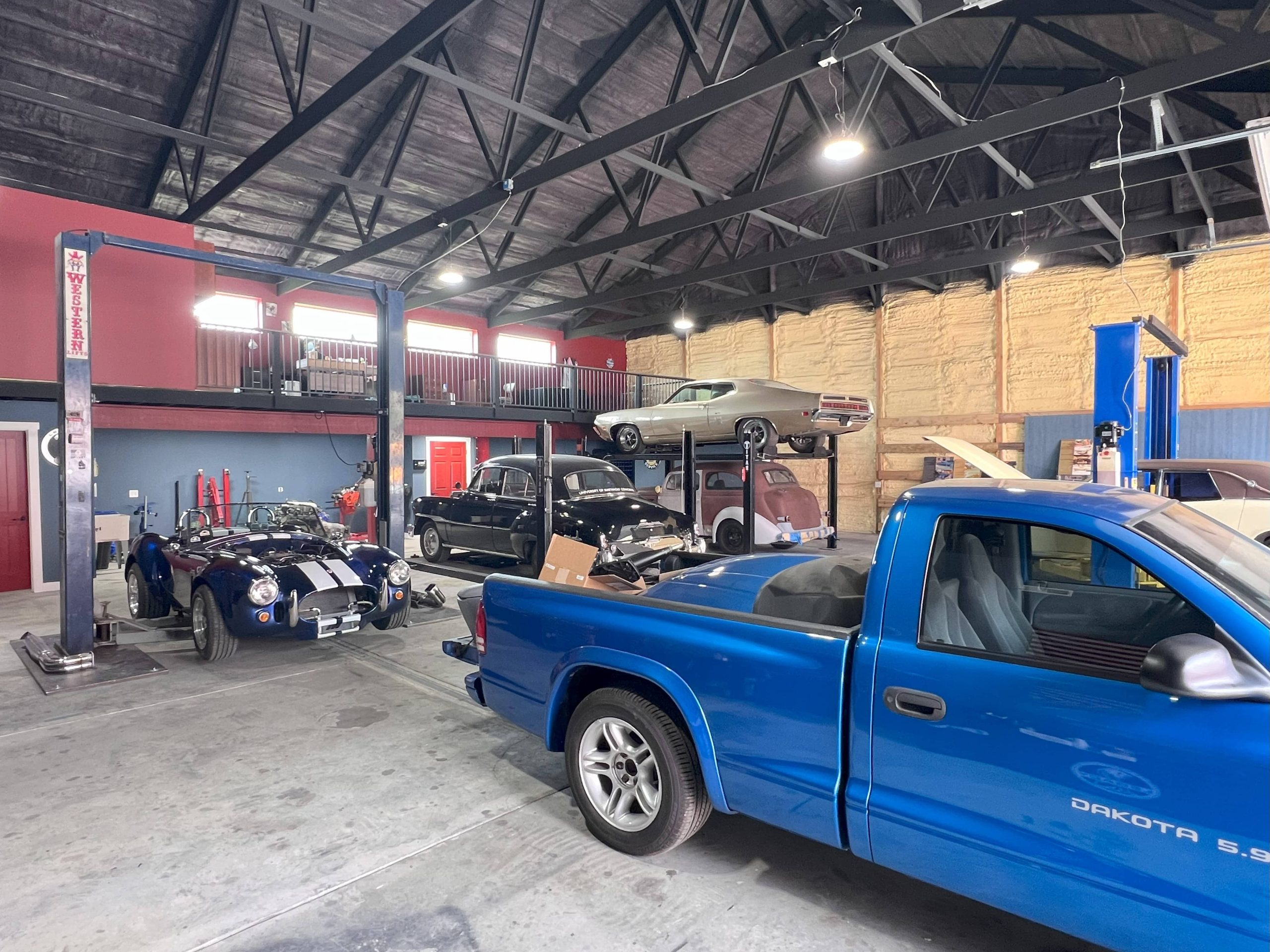 Interior of a custom auto garage featuring a blue Dodge Dakota, a Shelby-style Cobra, and several classic cars on lifts.