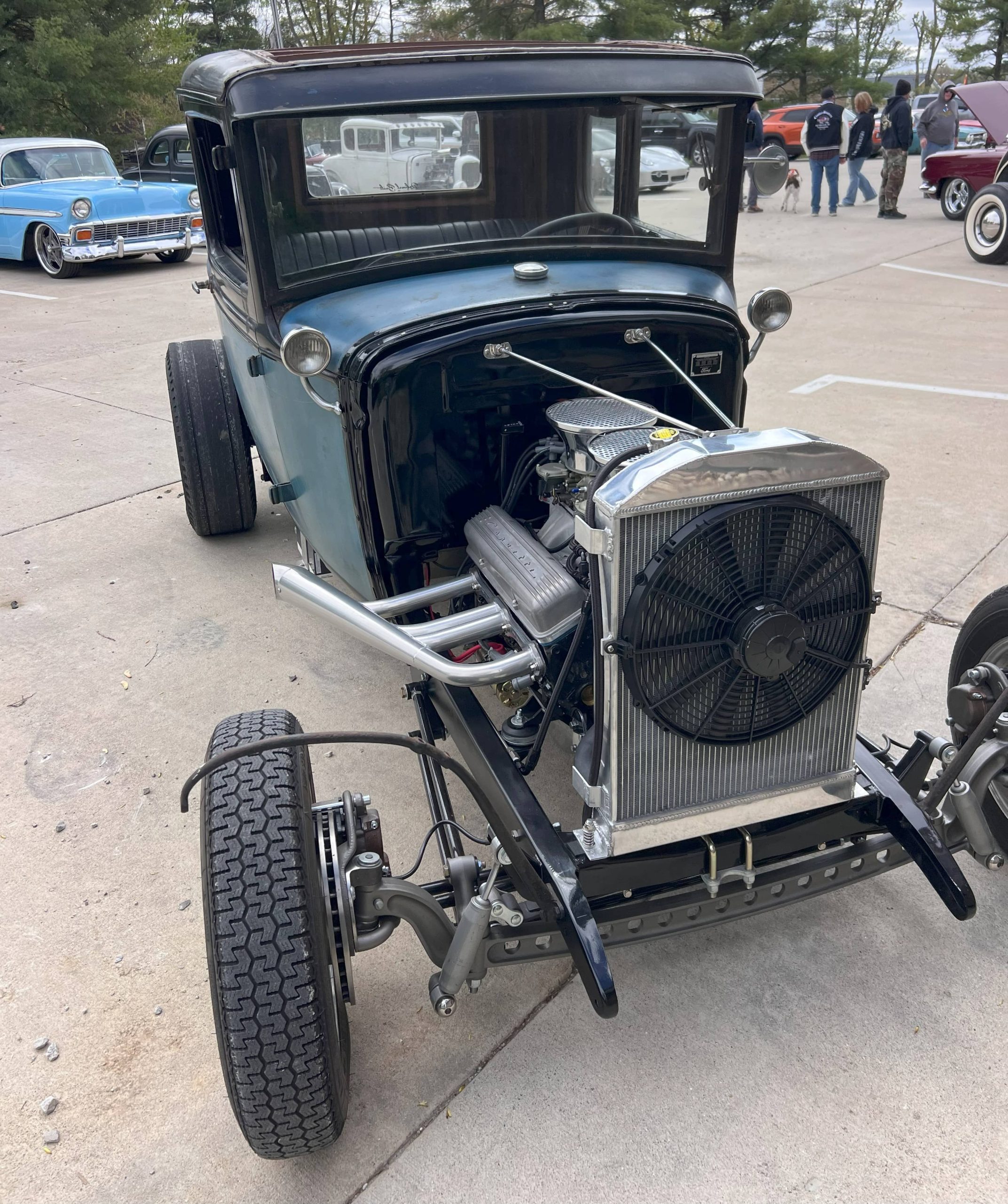 Front view of a vintage hot rod with an exposed engine and radiator, parked at a car show with other classic cars in the background.