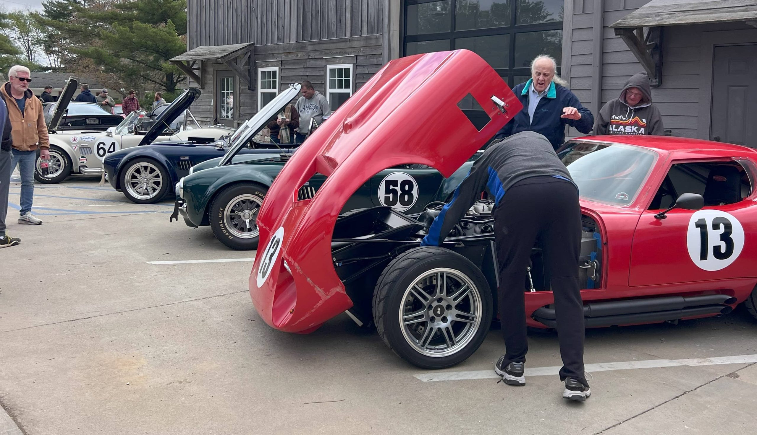 Red Daytona Coupe with number 13 and hood open, parked alongside Cobra-style roadsters at a car show with attendees observing.