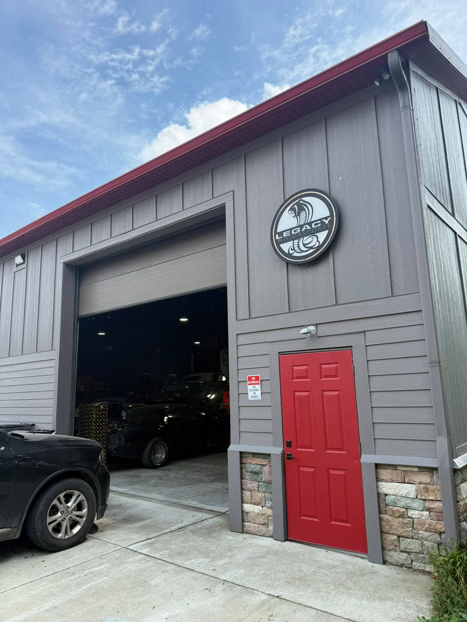 Exterior of the Legacy Motorsports garage with gray siding, an open service bay, and red entry door.