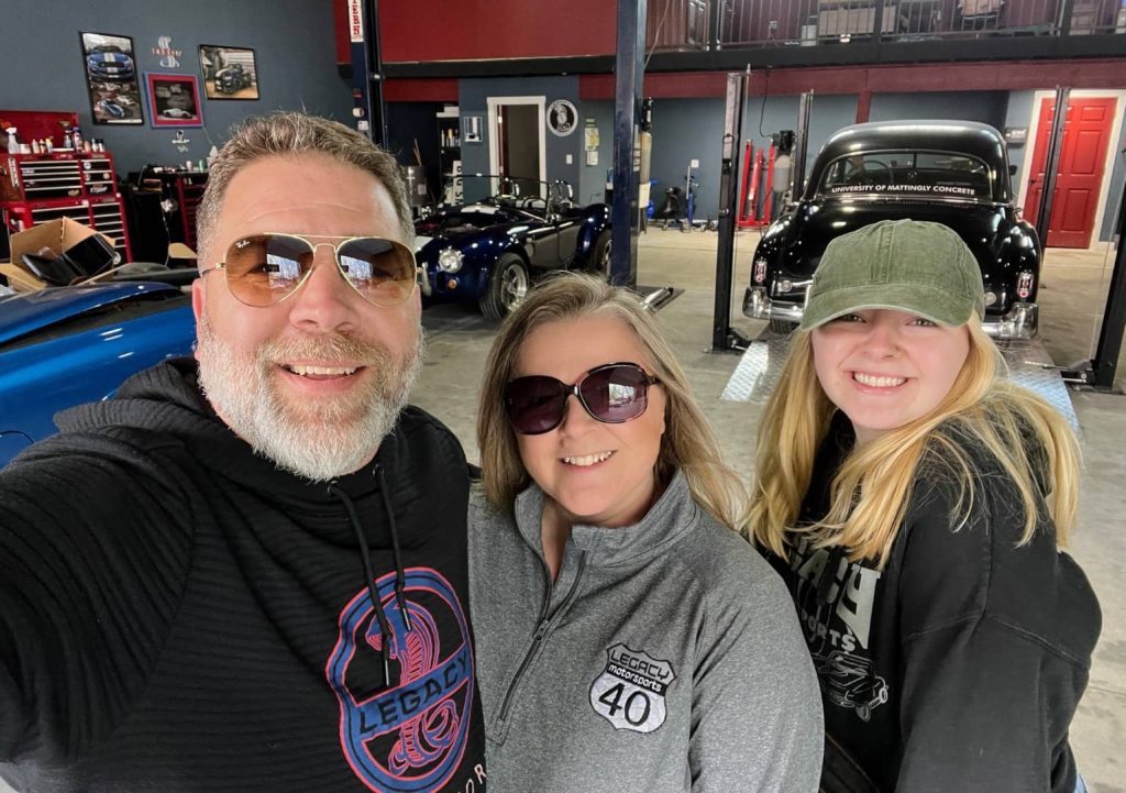 Three smiling Legacy Motorsports team members pose for a selfie inside the shop during Cars & Coffee Plainfield 2025, surrounded by classic cars and tools