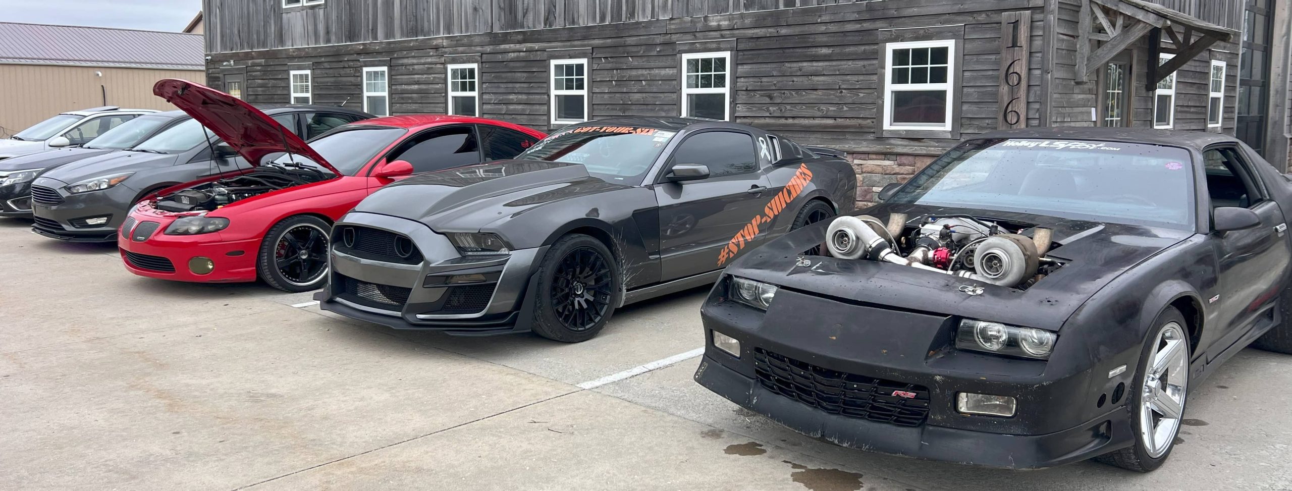 Row of performance and modified cars including a turbocharged Camaro, a widebody Mustang, and a red Pontiac GTO, parked outside a wooden building.
