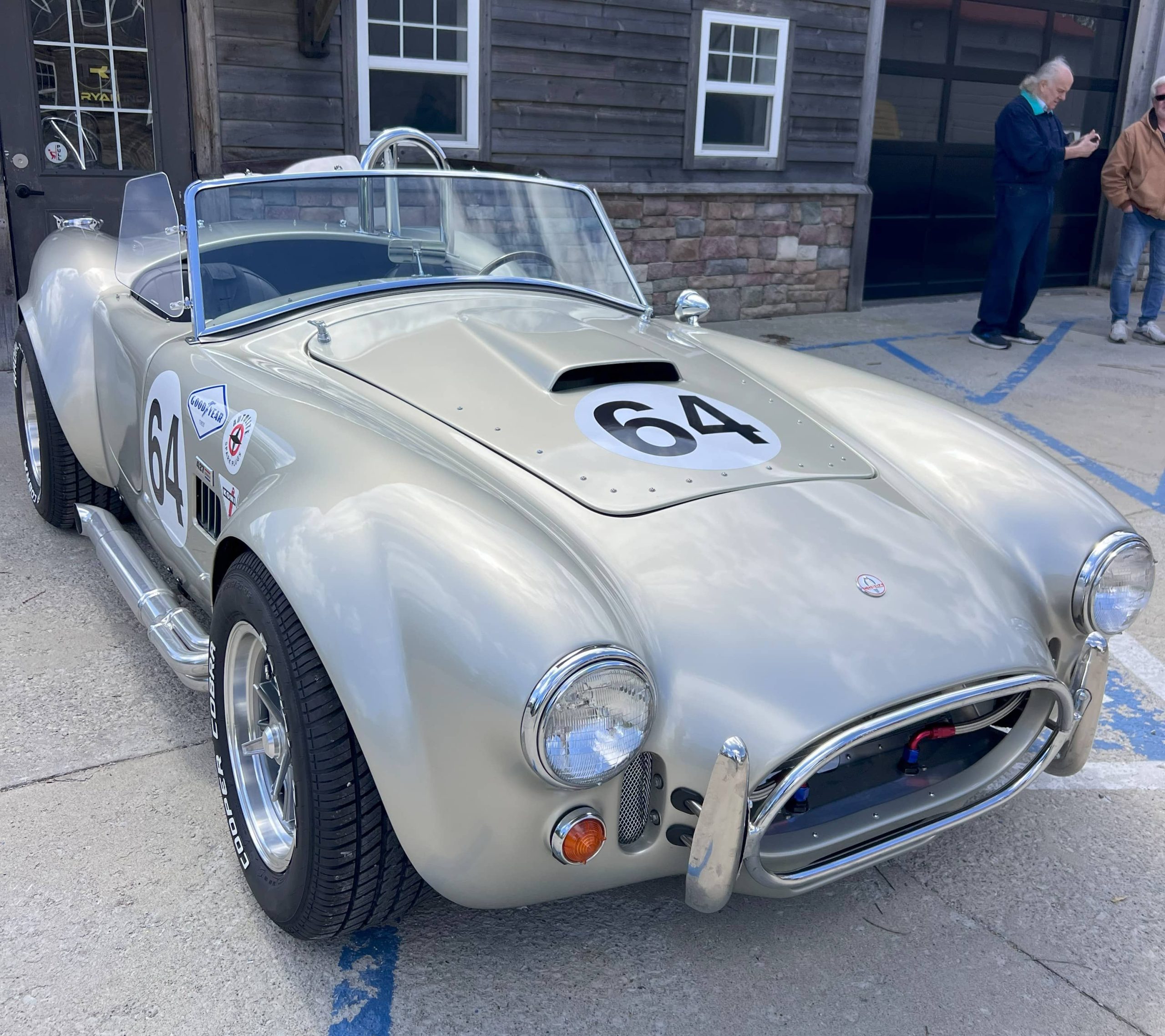 Silver Cobra-style race car with number 64 on the hood, parked at a car event with a stone and wood building in the background.