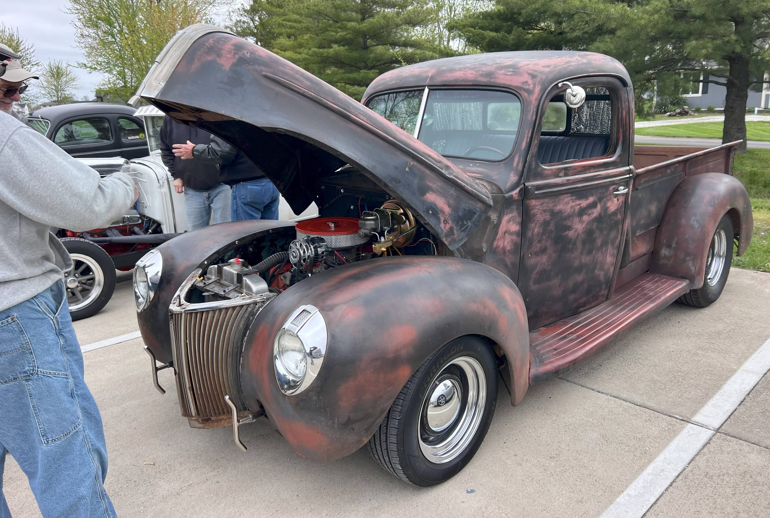 Rustic vintage pickup truck with a patina finish and open hood displaying the engine at a car show.
